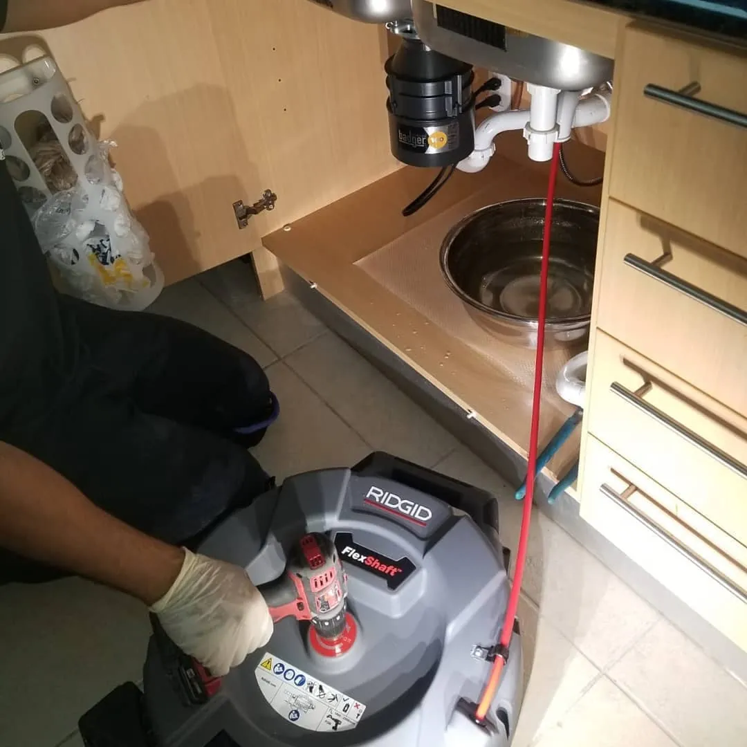 Gloved hands running a flexshaft machine whose red cable goes up into a kitchen sink drain