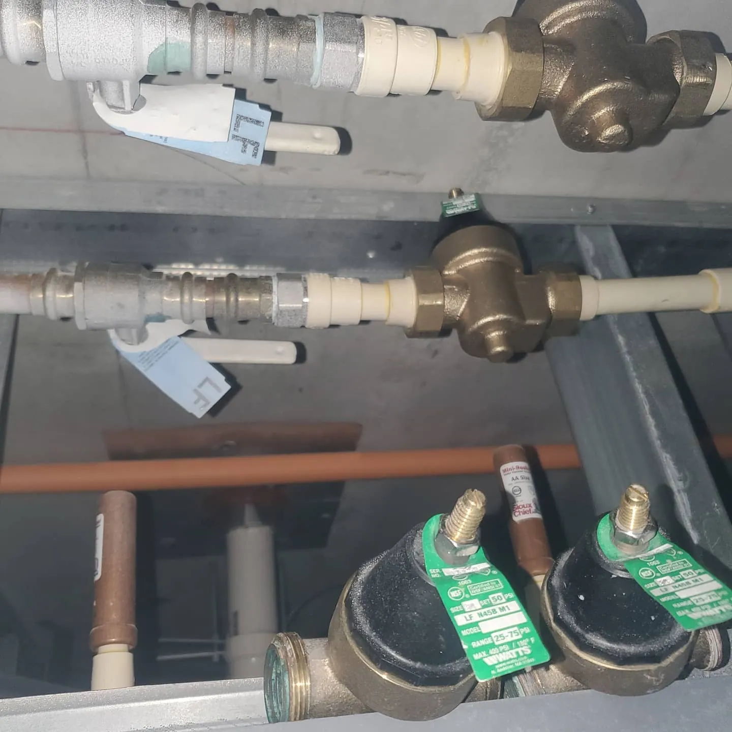 Two bronze pressure reducing valves with green tags installed on hot and cold lines overhead in a high-rise unit