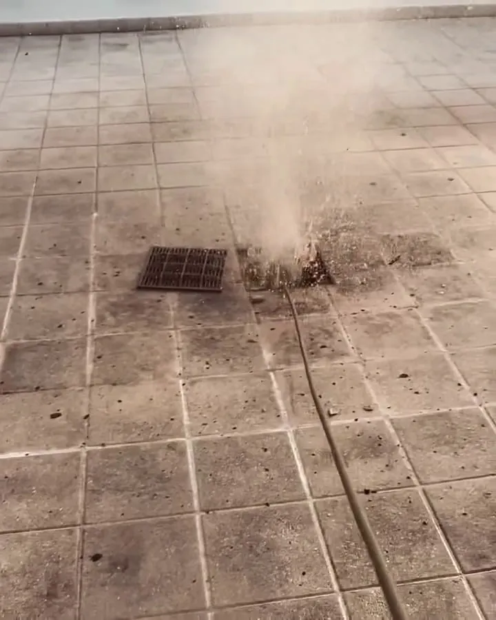 Water jet erupting from a rooftop drain, the jetting hose lying across wet tile toward the camera