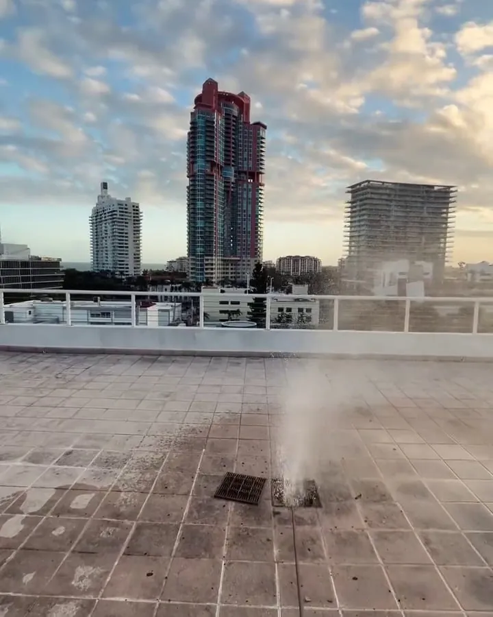 Jet of water shooting up from a rooftop drain on a high-rise, with the skyline behind