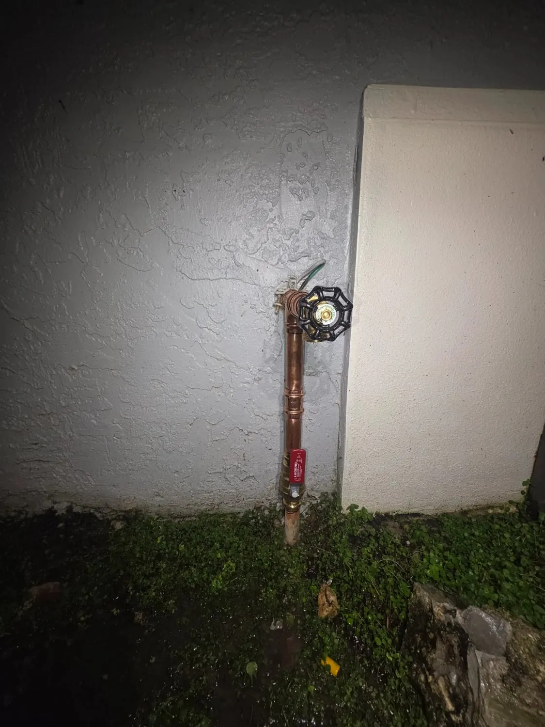 New copper stub with a red ball valve and a new brass hose bib on a gray stucco wall at night
