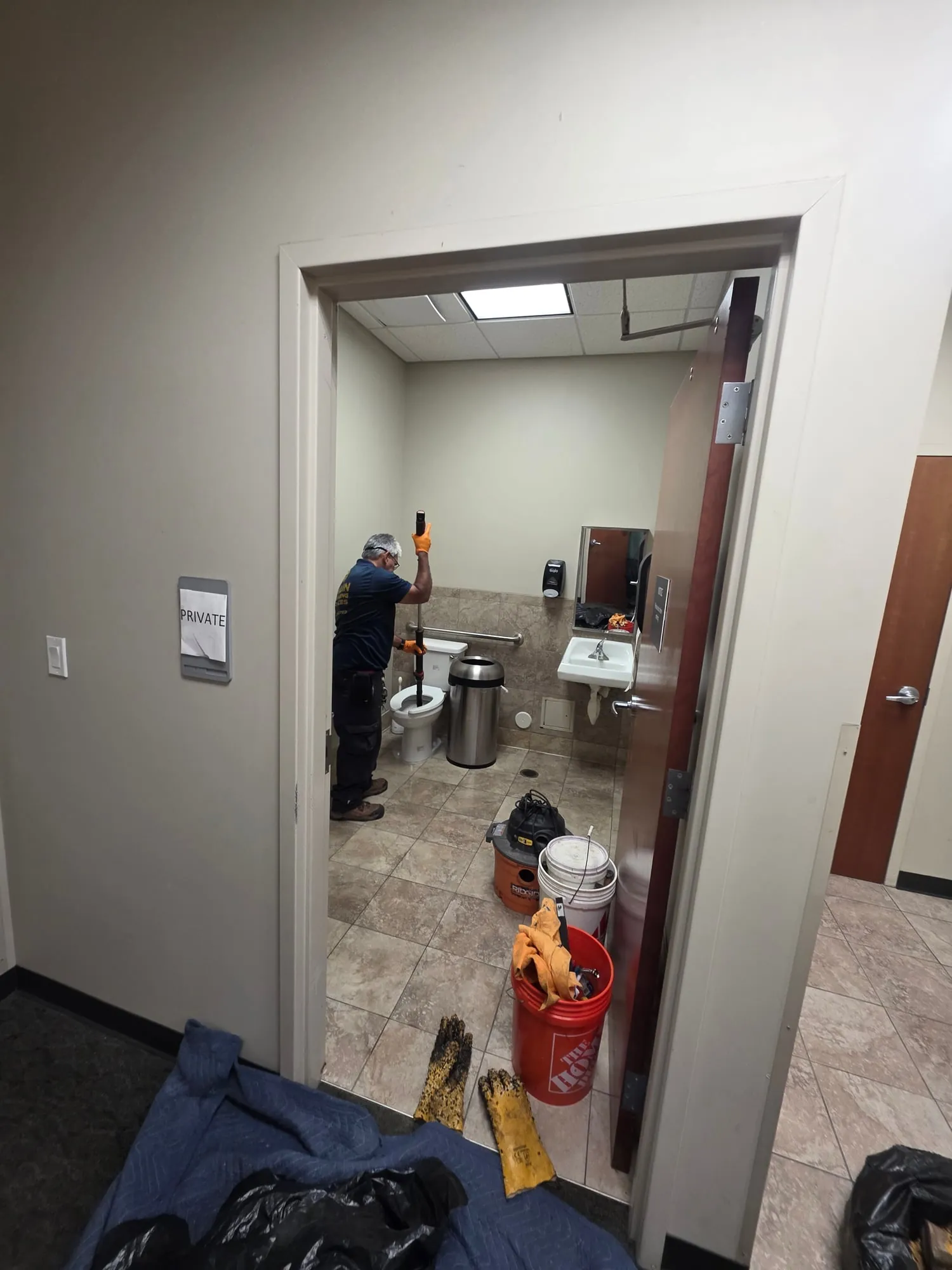 Plumber working at a clogged toilet in an office restroom, with a wet vacuum and buckets in the doorway