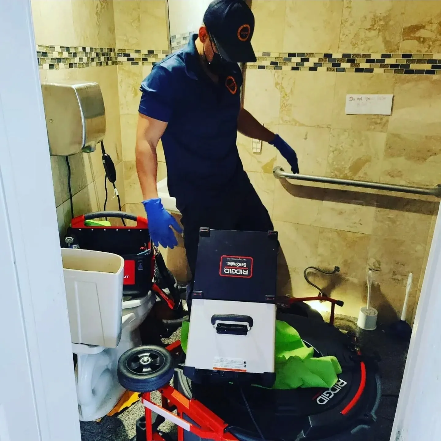 Plumber in a commercial restroom with a RIDGID SeeSnake sewer camera reel and monitor set up on the floor