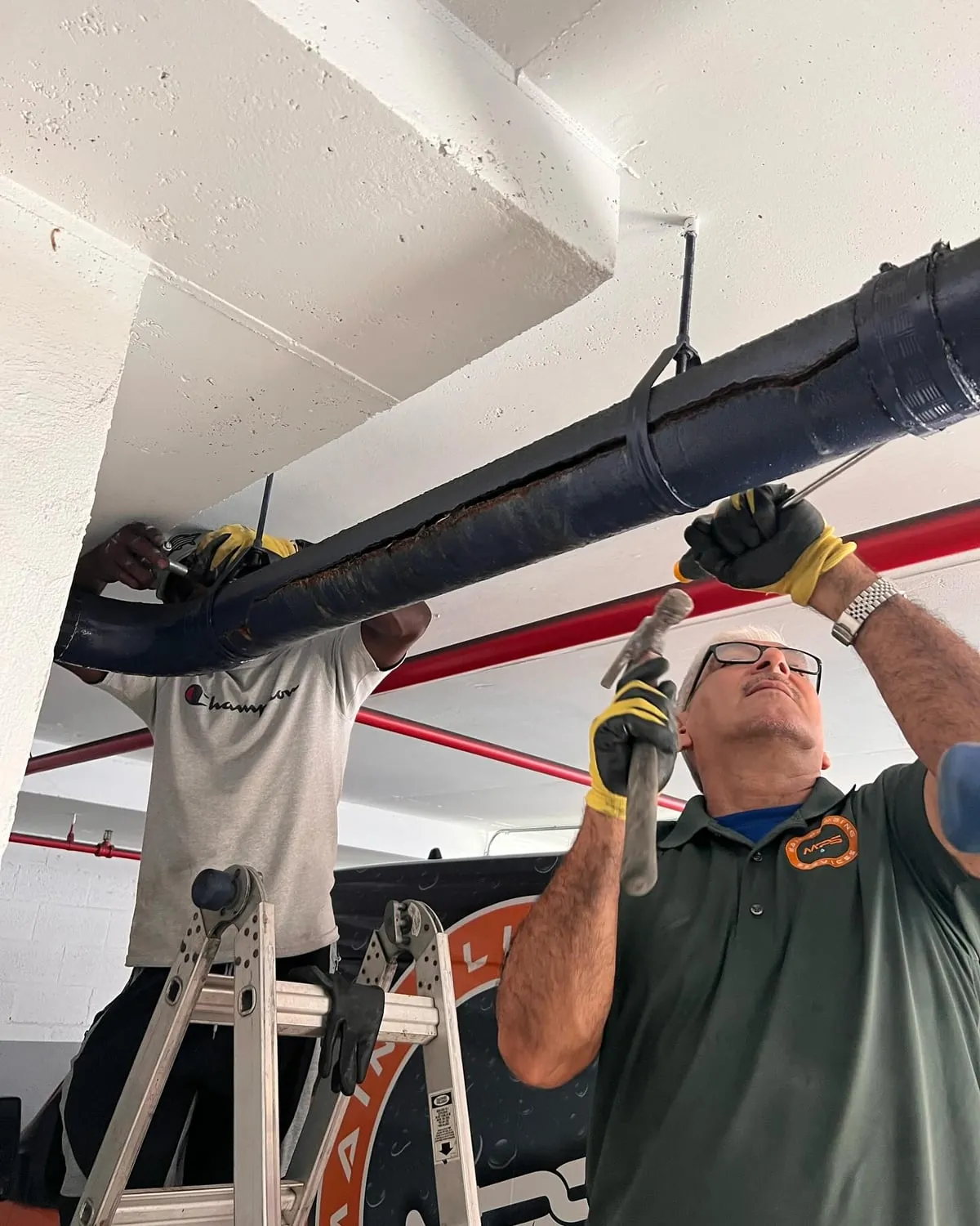 Two plumbers on a ladder replacing an overhead cast iron drain line in a parking garage