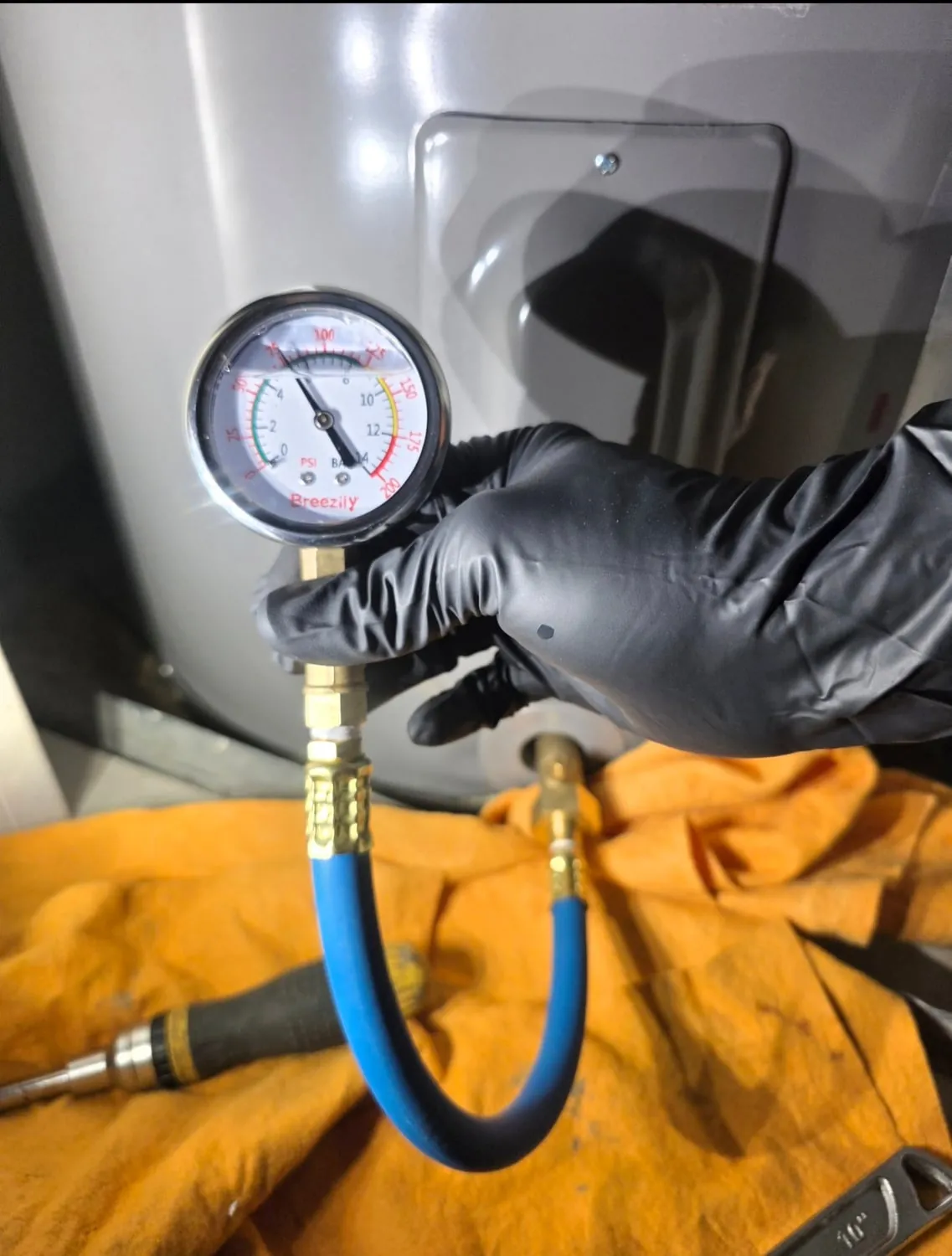 A water pressure gauge on a short blue hose, connected to a fitting at the bottom of a water heater