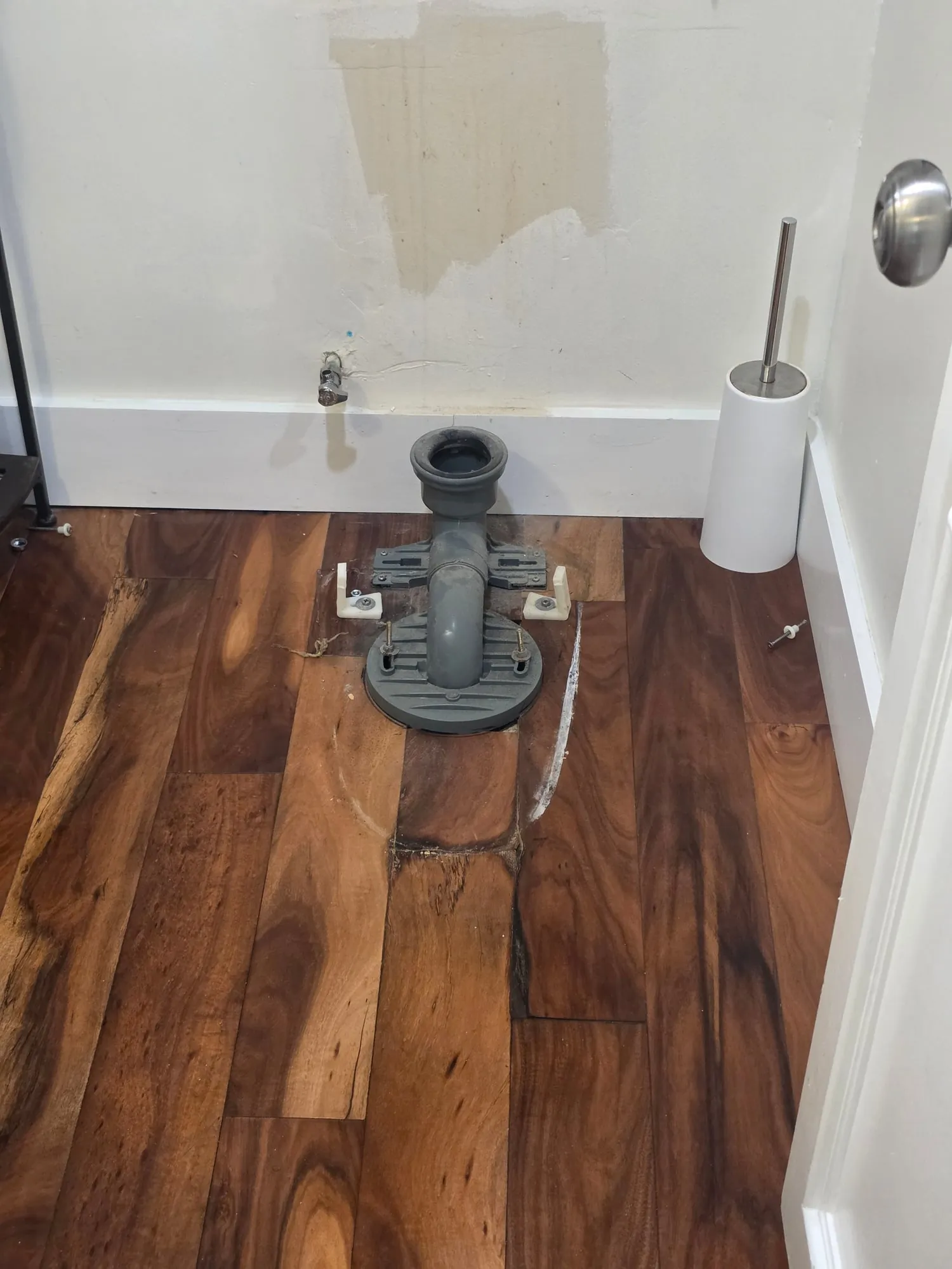Gray closet flange and bend exposed in a hardwood bathroom floor after the old toilet was removed