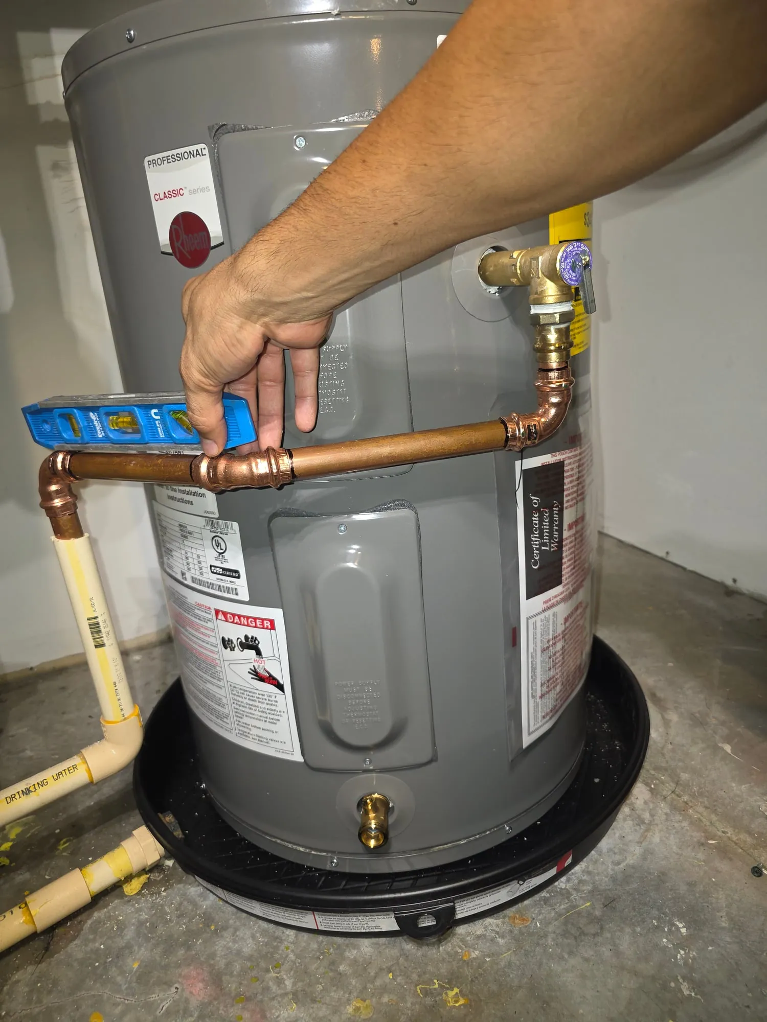 Hand holding a level on a new copper hot-water line above an electric water heater with a relief valve and drain pan