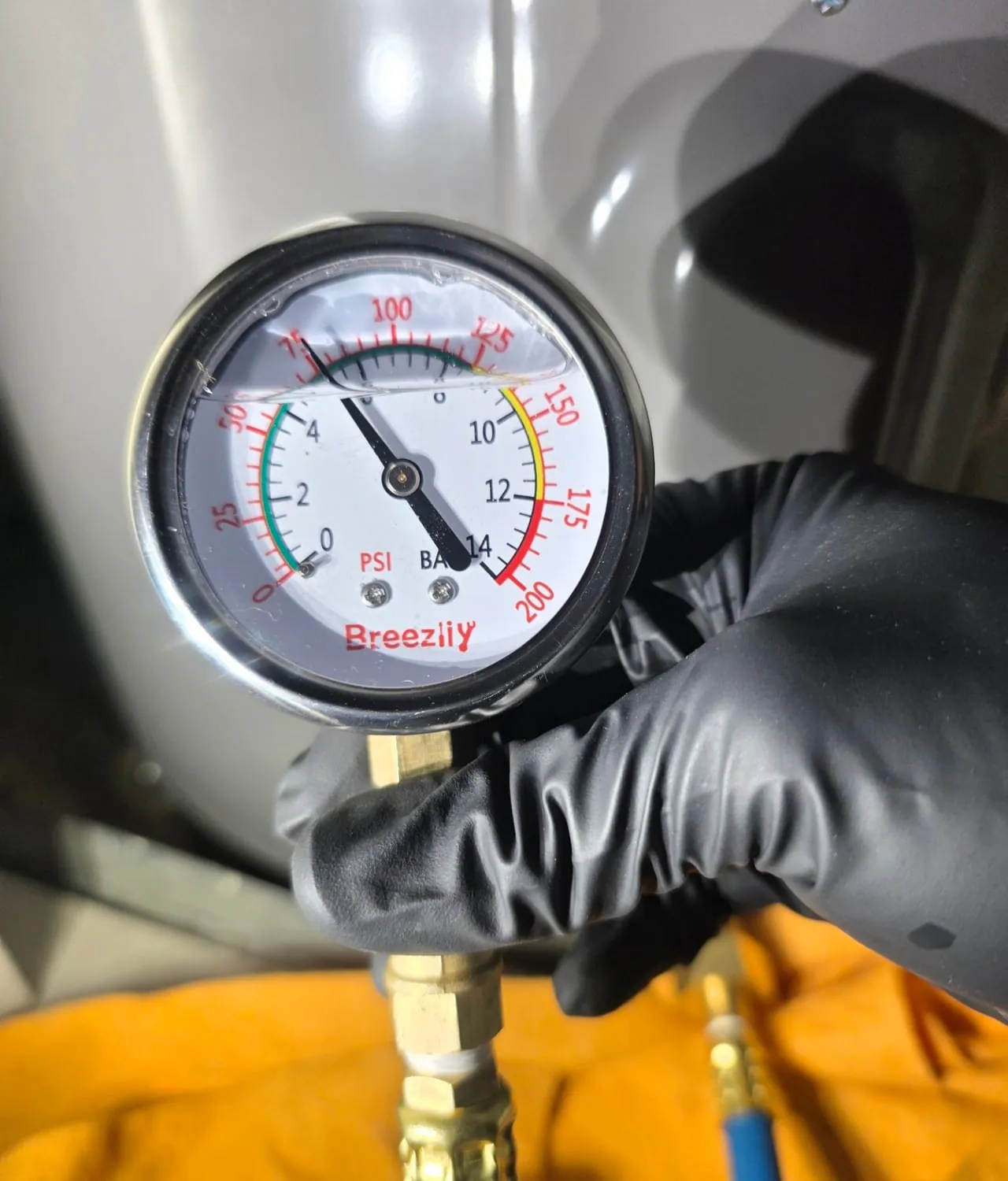 A gloved hand holding a water pressure gauge in front of a water heater tank